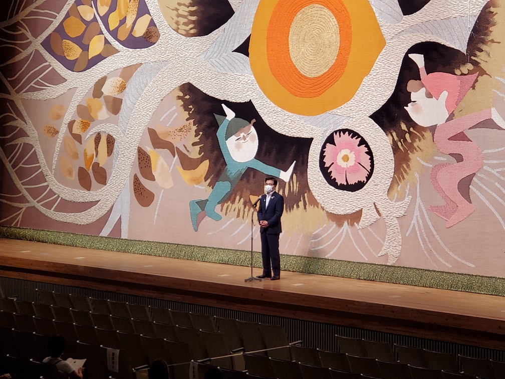 鮮やかな色と抽象的なデザインの緞帳が下りた舞台中央に市長が立ち、マイクでスピーチをしている写真