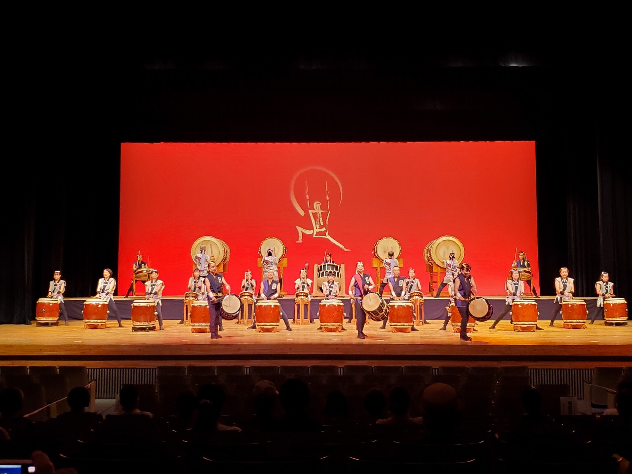 背後に赤い照明に浮かび上がる人型のシンボルマークが描かれている舞台上に、大きな和太鼓が多数並べられ、白い衣装を着た複数の演奏者が力強く太鼓を叩いている写真