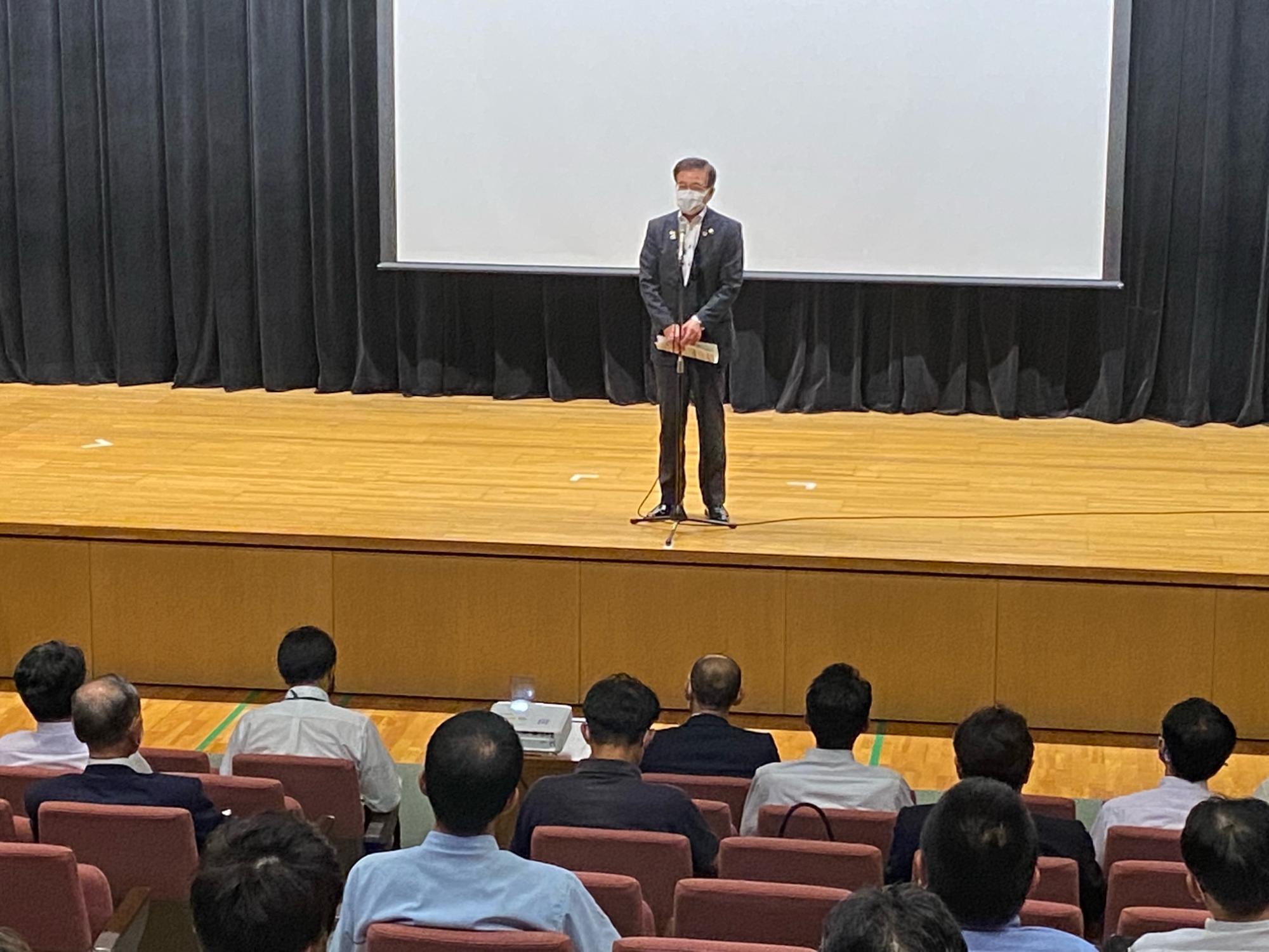 広いステージ上で、スーツ姿の市長がスタンドマイクを前に立ち、多くの聴衆が席に座っている写真