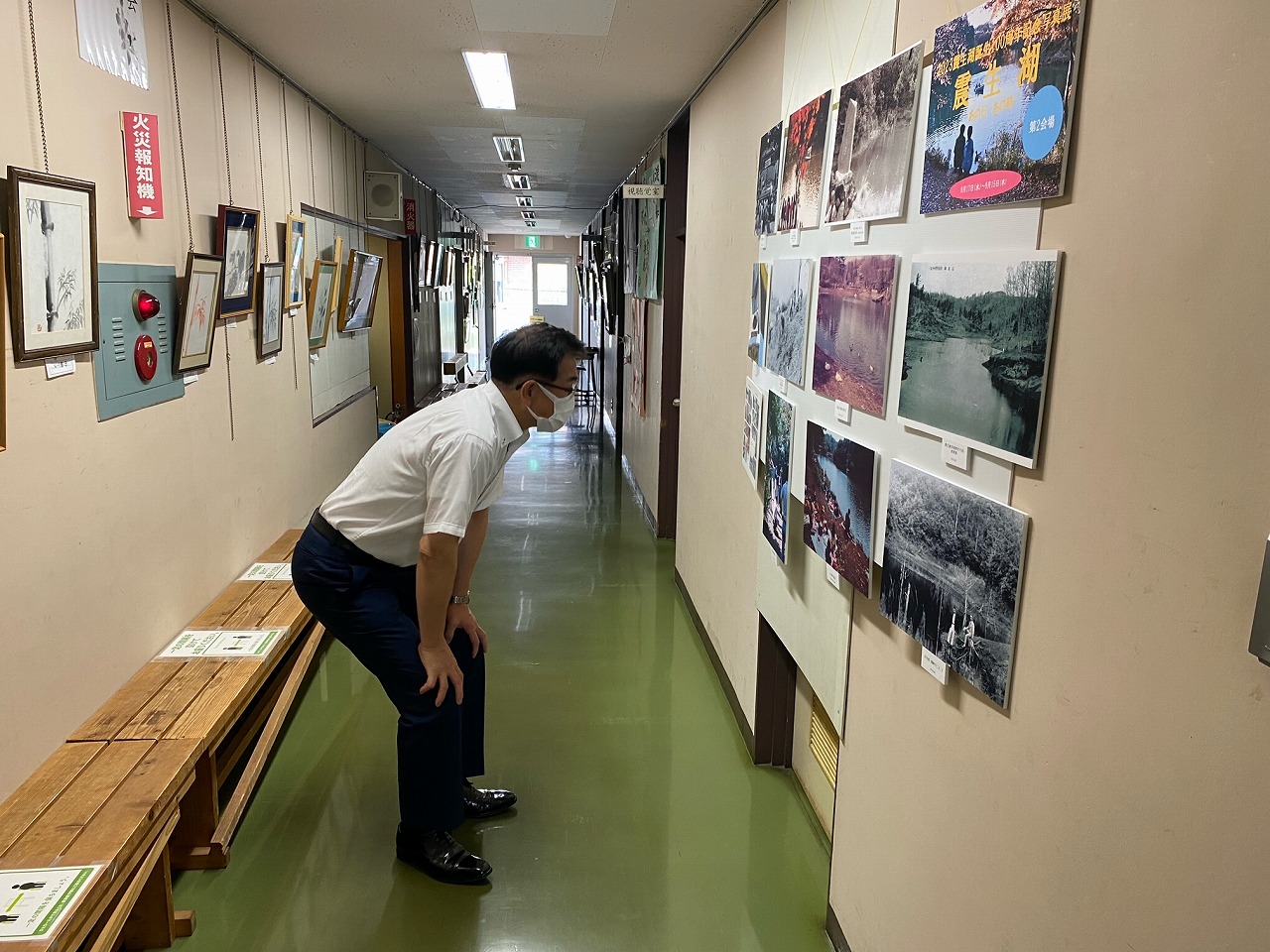 壁にたくさんの写真が展示された廊下で、マスクを着用した市長が腰をかがめて熱心に鑑賞している様子の写真