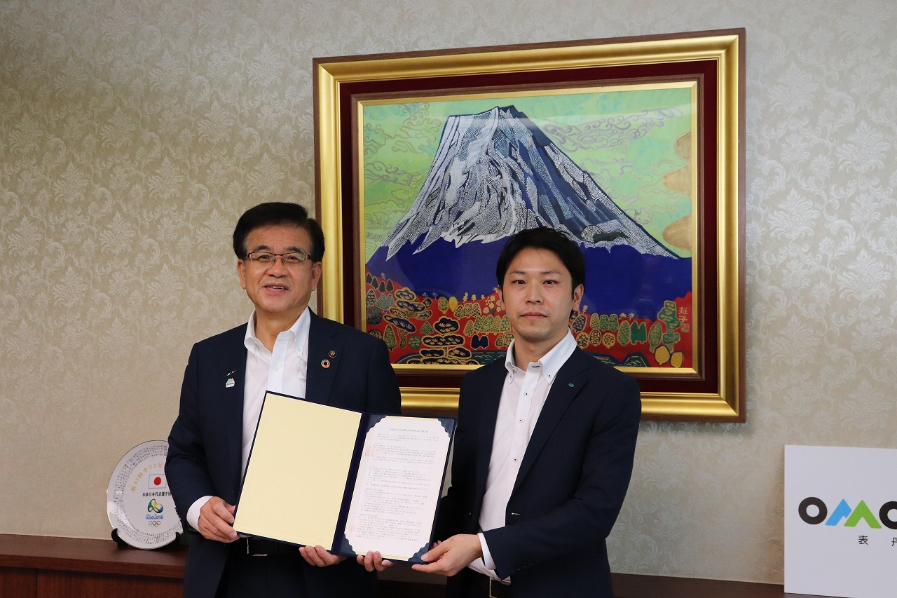 富士山の絵画が飾られた室内で、2人のスーツ姿の市長と男性が、協定書を広げている写真