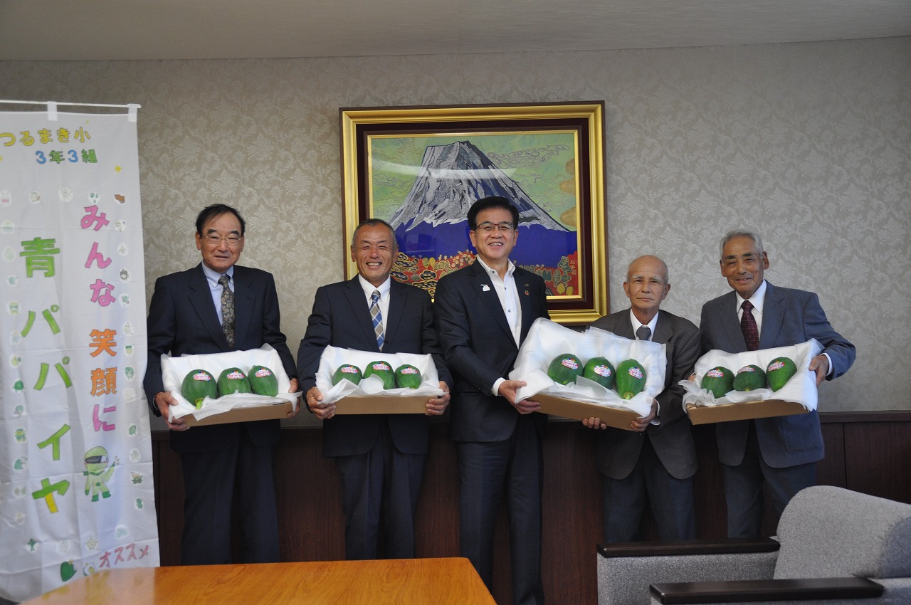 スーツ姿の男性5名が青パパイアが3個ずつはいった箱をそれぞれが持ち笑顔で記念撮影をしている写真