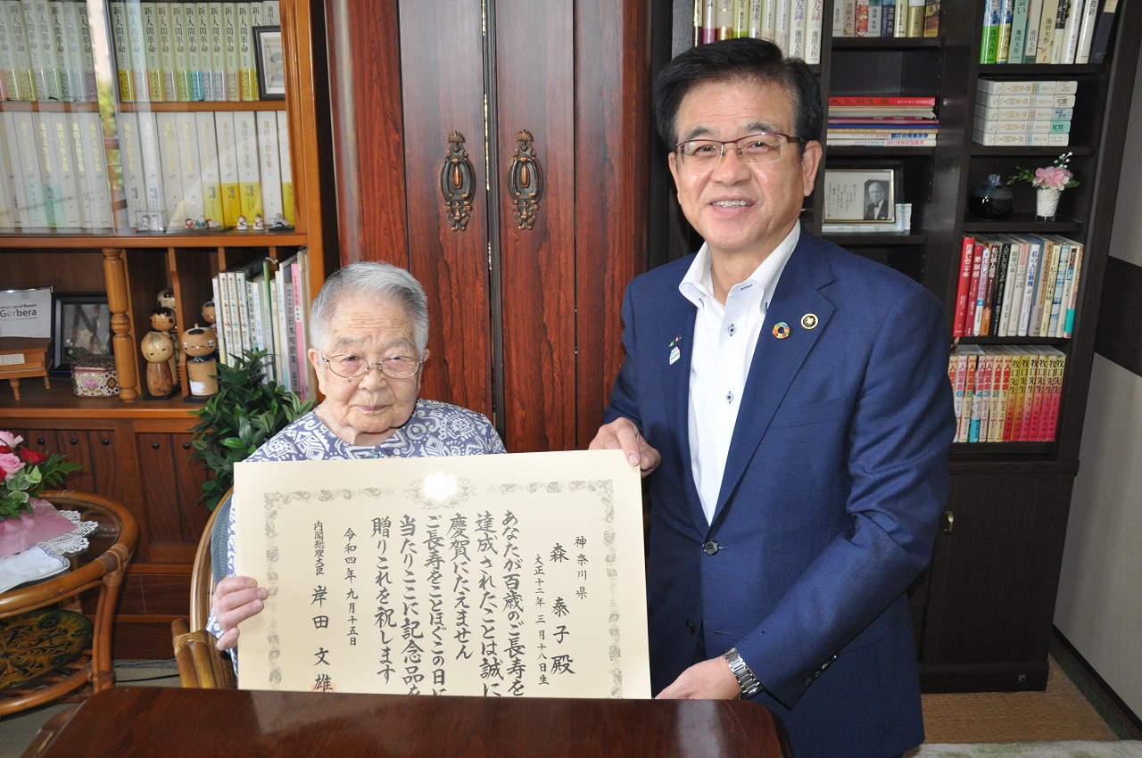 市長が座って賞状をもつ森泰子さんと並んで記念撮影をしている写真