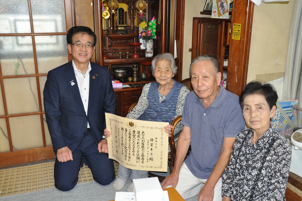 自宅の和室で、スーツ姿の男性が感謝状を広げた諸星キミさん夫婦と女性と共に座り、カメラに向かって笑顔で写っている記念写真