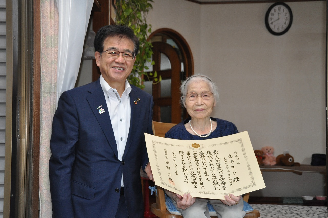 眼鏡をかけたスーツ姿の男性が感謝状を持つ金澤ミツさんの横に立っており、二人ともカメラに向かって笑顔を見せている室内の写真