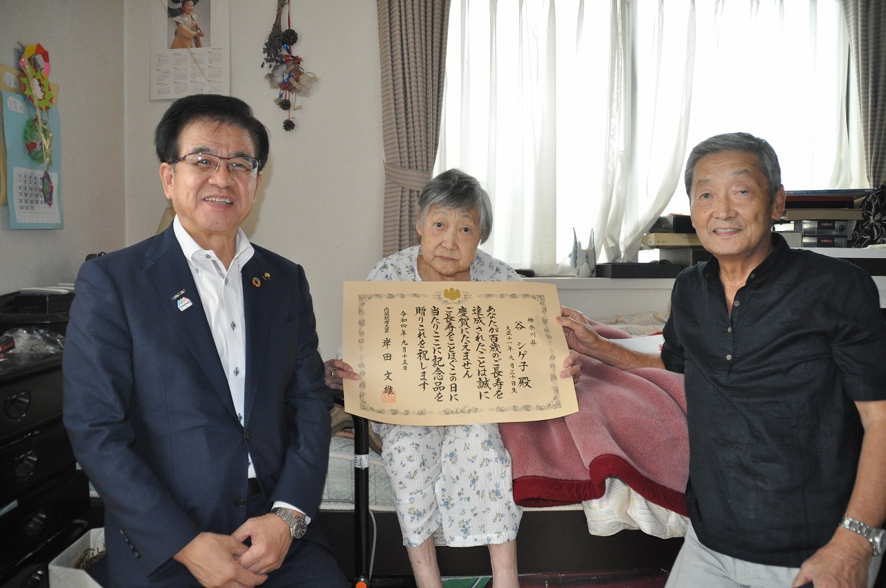 ベッドに腰かけた谷シゲ子さんを中心に左にスーツ姿の市長、右に一緒に賞状に手をそえている男性の3人でカメラに向かって笑顔を見せている室内の写