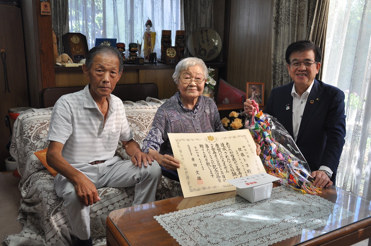 テーブルには記念品が置かれソファーに賞状を見せながら座る佐藤トヨさんと男性、スーツ姿の市長が折り鶴をもって記念撮影をしている写真