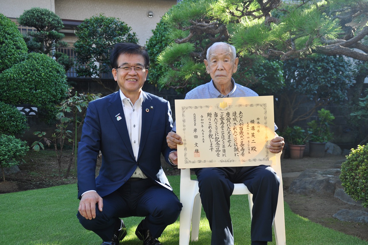 庭先でスーツ姿の男性がしゃがみ、白い椅子に座った横溝英二さんが賞状をカメラに向かって掲げており、二人が笑顔を見せている屋外での記念写真