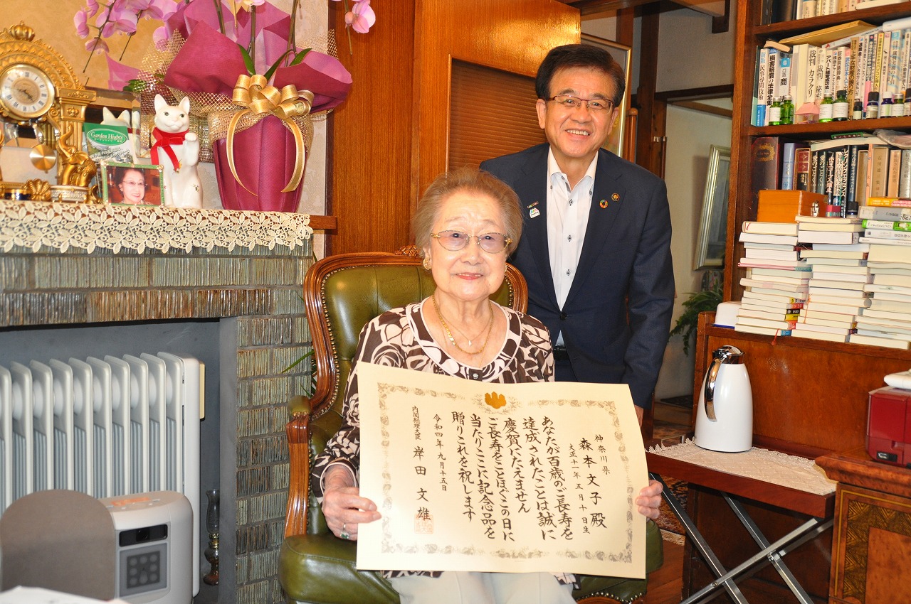 室内の椅子に座った森本文子さんが賞状をカメラに見せながらその後ろでスーツ姿の市長が並んで記念撮影をしている写真