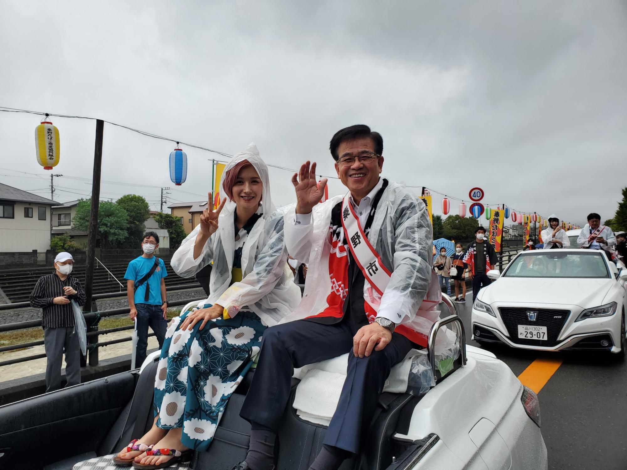 和やかな雰囲気のパレードで、雨合羽を着た男女がオープンカーに乗って手を振り、沿道に並んだ観客や提灯に彩られた街並みの中を進んでいる様子の写真