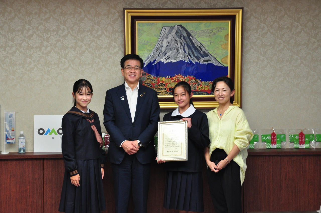 市長の両隣に制服姿の女子中学生が並び、左隣の女の子が賞状を見せながら隣に立つ女性4名で笑顔で記念撮影をしている写真