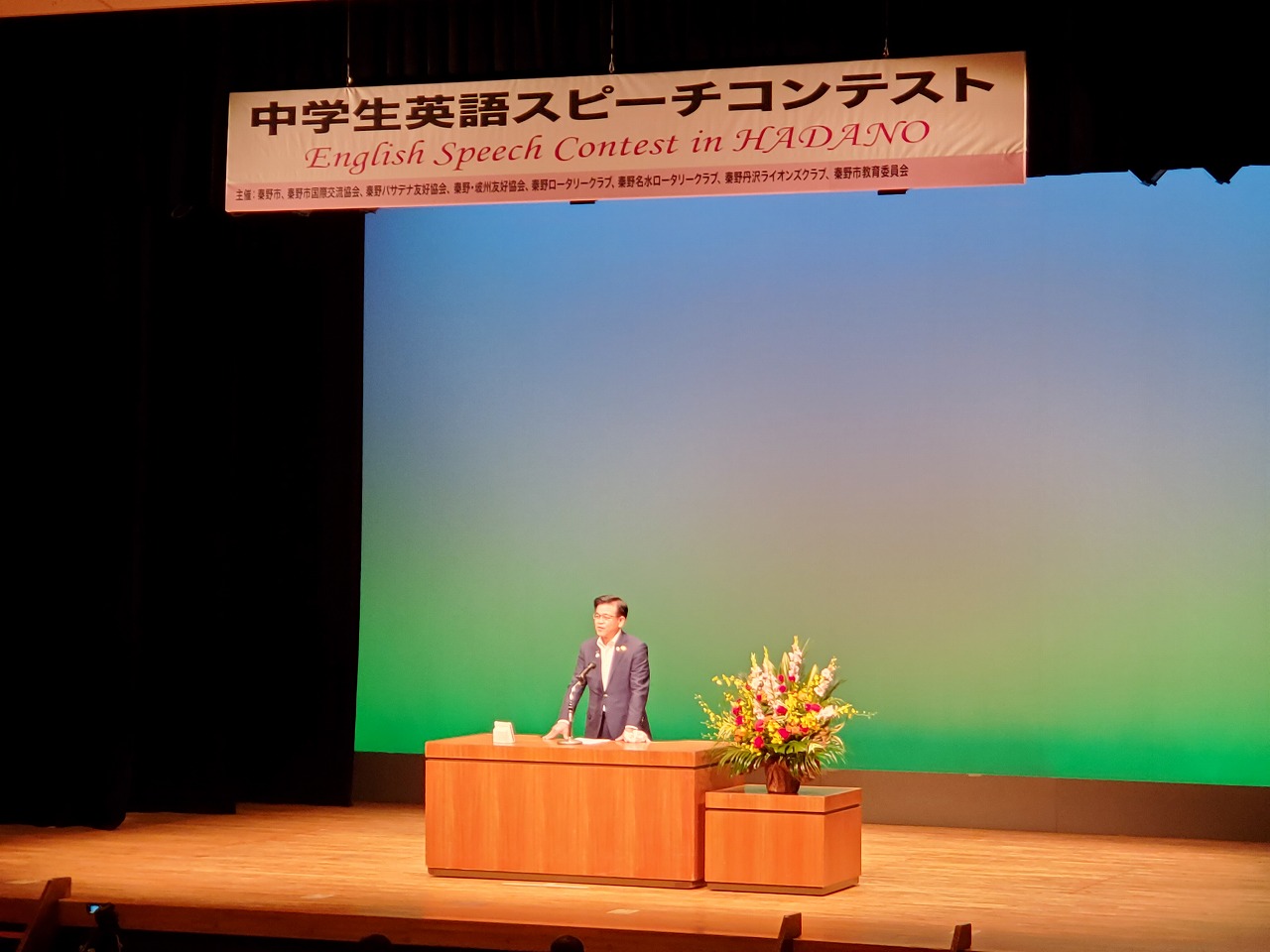 木製の演台に立つ男性が聴衆に向かって話しており、ステージ上部には「中学生英語スピーチコンテスト」と書かれた横断幕が掲げられている写真