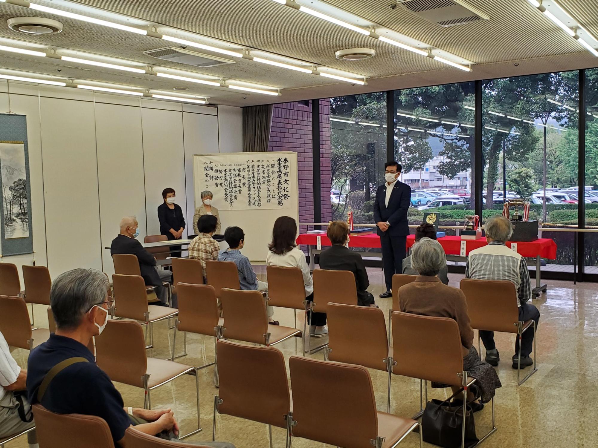 和やかな雰囲気の中、マスクを着用した男性が椅子に座る聴衆に話しかけており、背後には賞状やトロフィーが並んだテーブルがある室内の写真