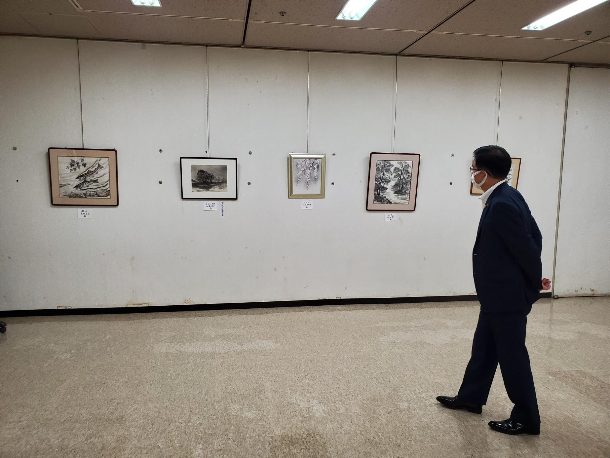 ギャラリーのような白い壁に水墨画や日本画が飾られており、マスクをつけたスーツ姿の男性が鑑賞している様子の写真