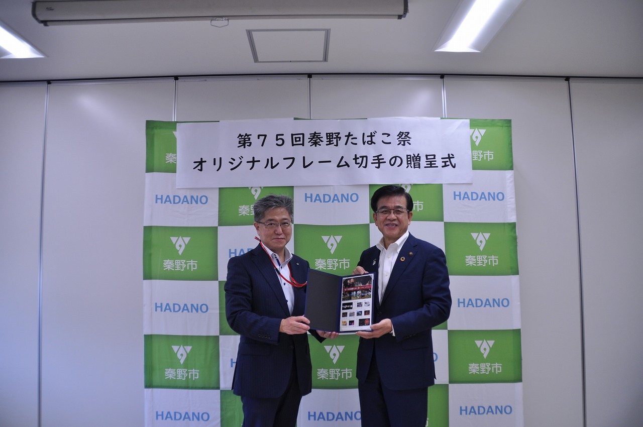 緑と白の市松模様のパネルを背景に、スーツ姿の男性二人が笑顔でオリジナルフレーム切手を持ち、カメラにポーズをとっている写真