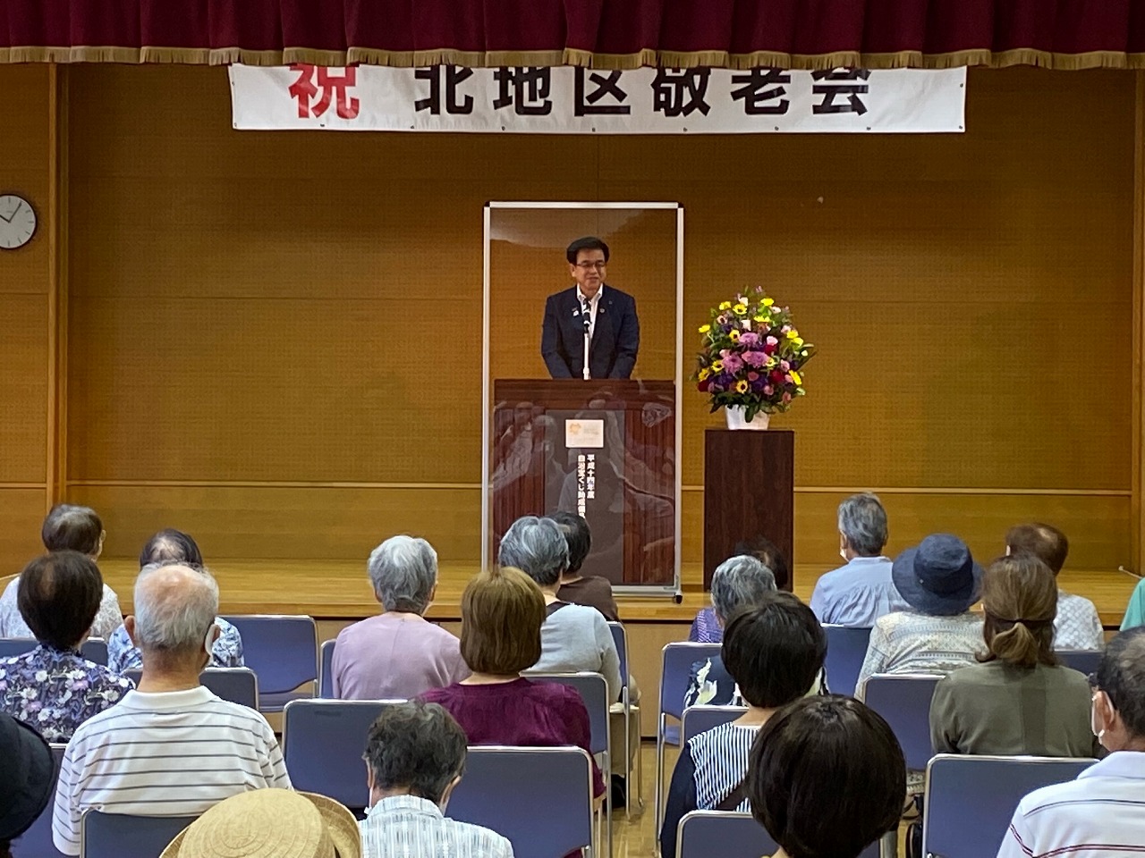 檀上でマイクに向かって話すスーツ姿の男性を、会場の椅子に座る多くの高齢者が後ろ姿で見守っており、ステージ上部には「祝 北地区敬老会」と書かれた横断幕がある写真