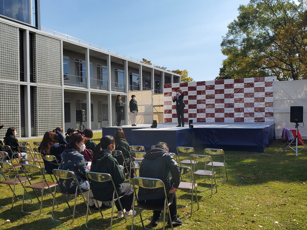 天候の良い屋外の広場で、ステージに立つ男性が話をし、椅子に座り話を聞いている方を後方から写した写真