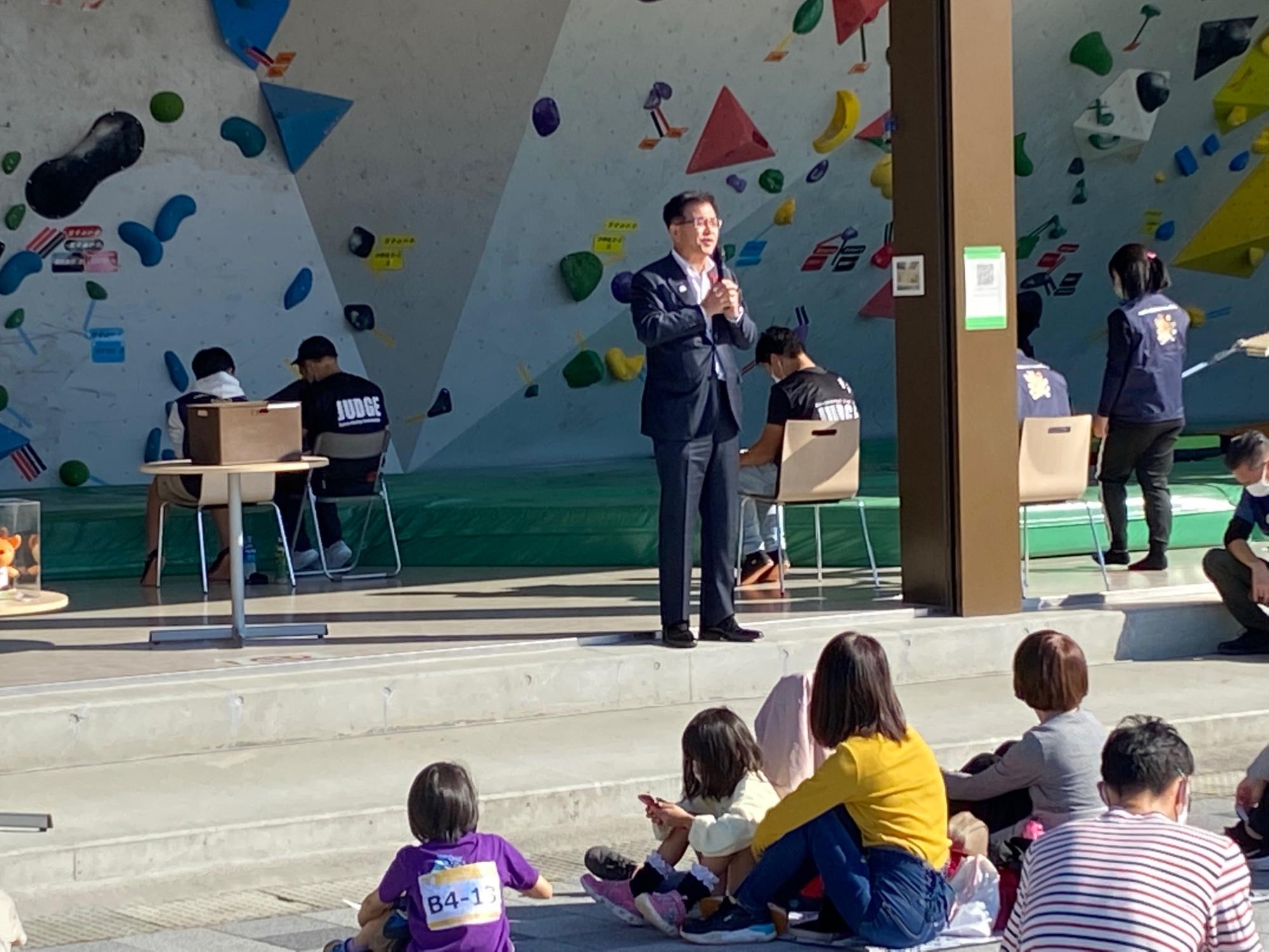 ボルダー壁の前でスーツ姿の男性がマイクを持って話しており、その前で複数の方々が座って話を聞いている写真