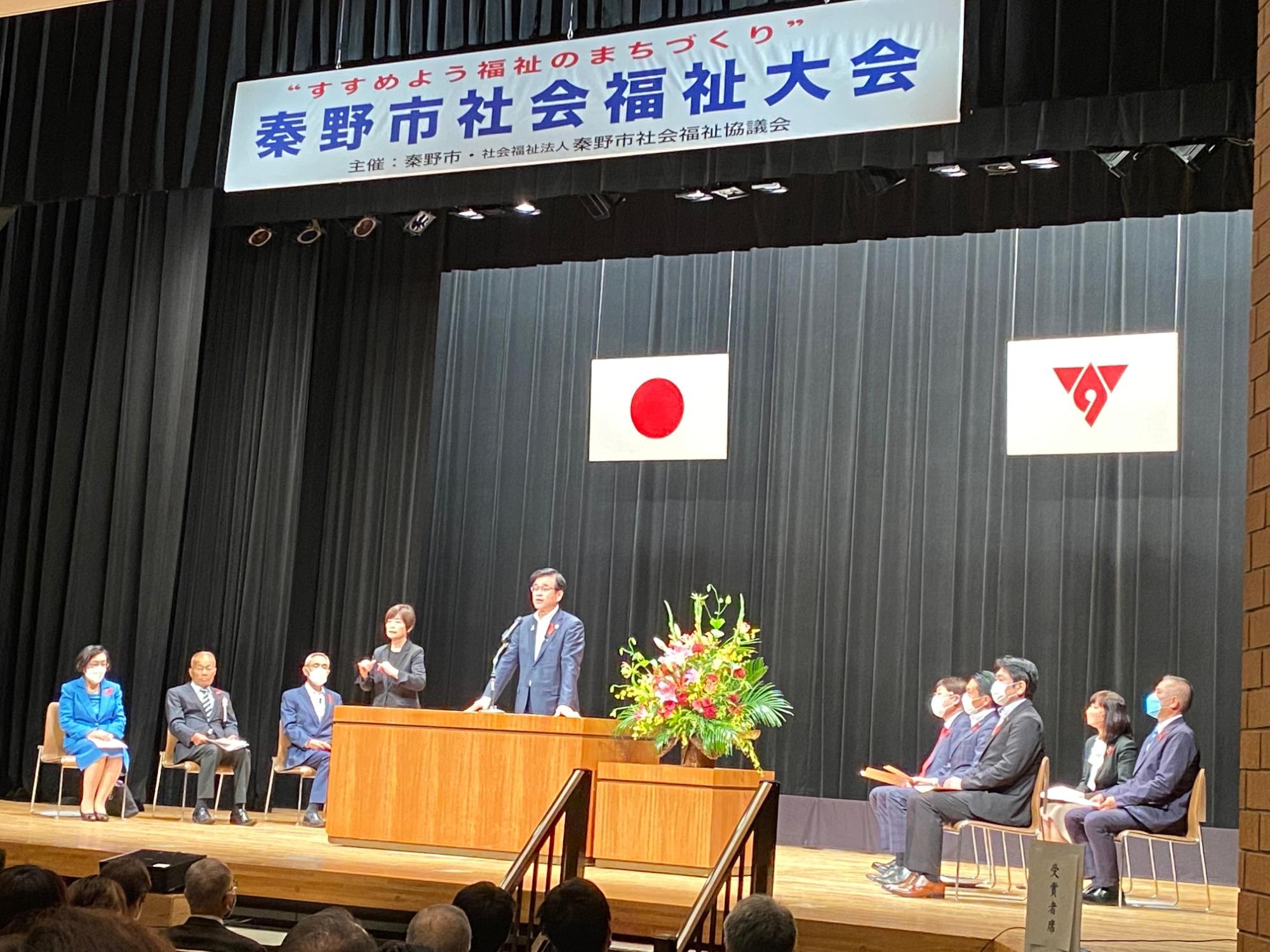 壇上でスーツ姿の男性が演台に立って話しており、背景には「秦野市社会福祉大会」と書かれた横断幕が掲げられ、複数の出席者が左右に着席している式典会場の写真