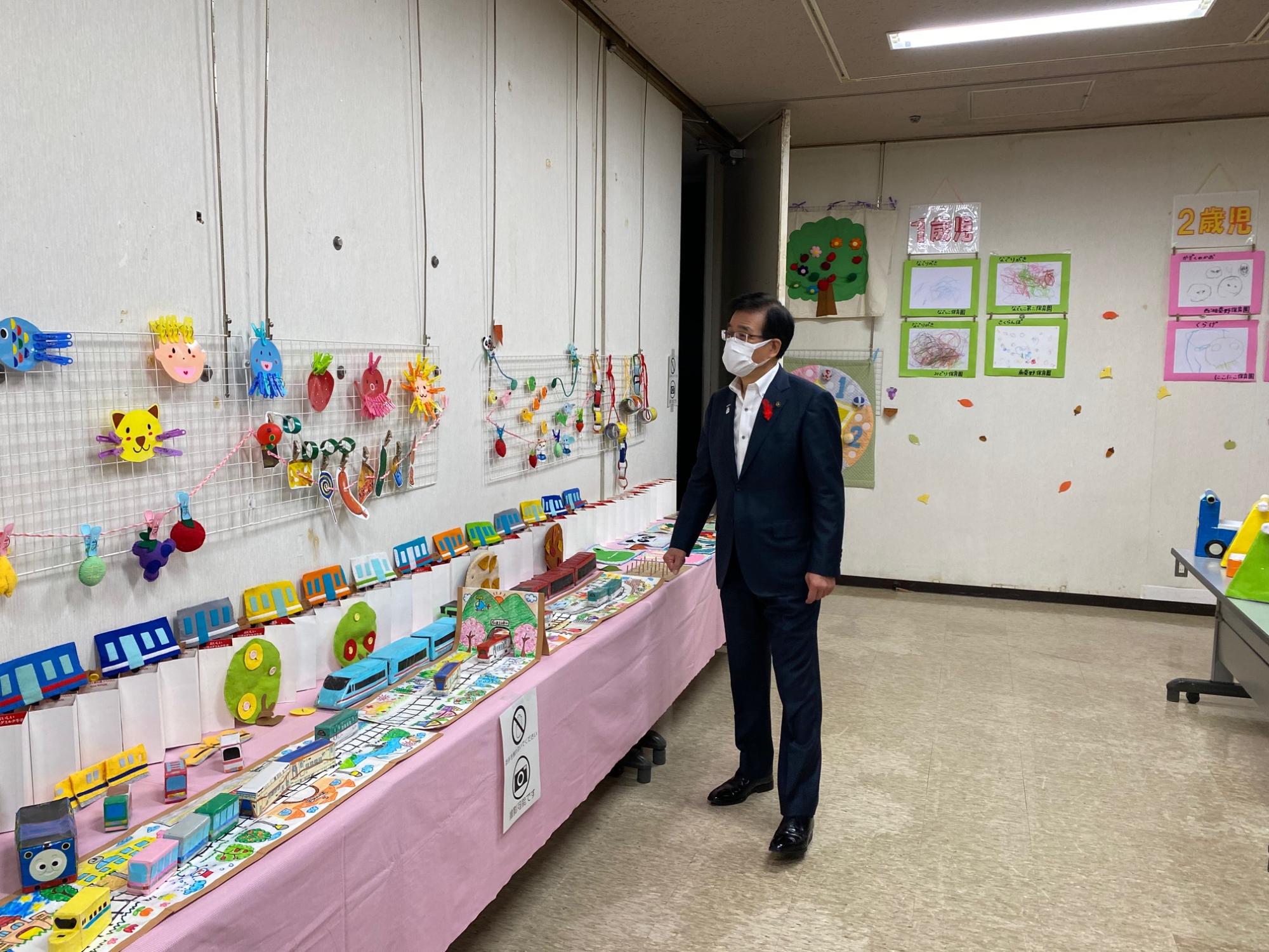 スーツ姿でマスクをした男性が、子どもたちが作成した電車や果物などのカラフルな工作作品が並べられた室内展示を見ている様子の写真