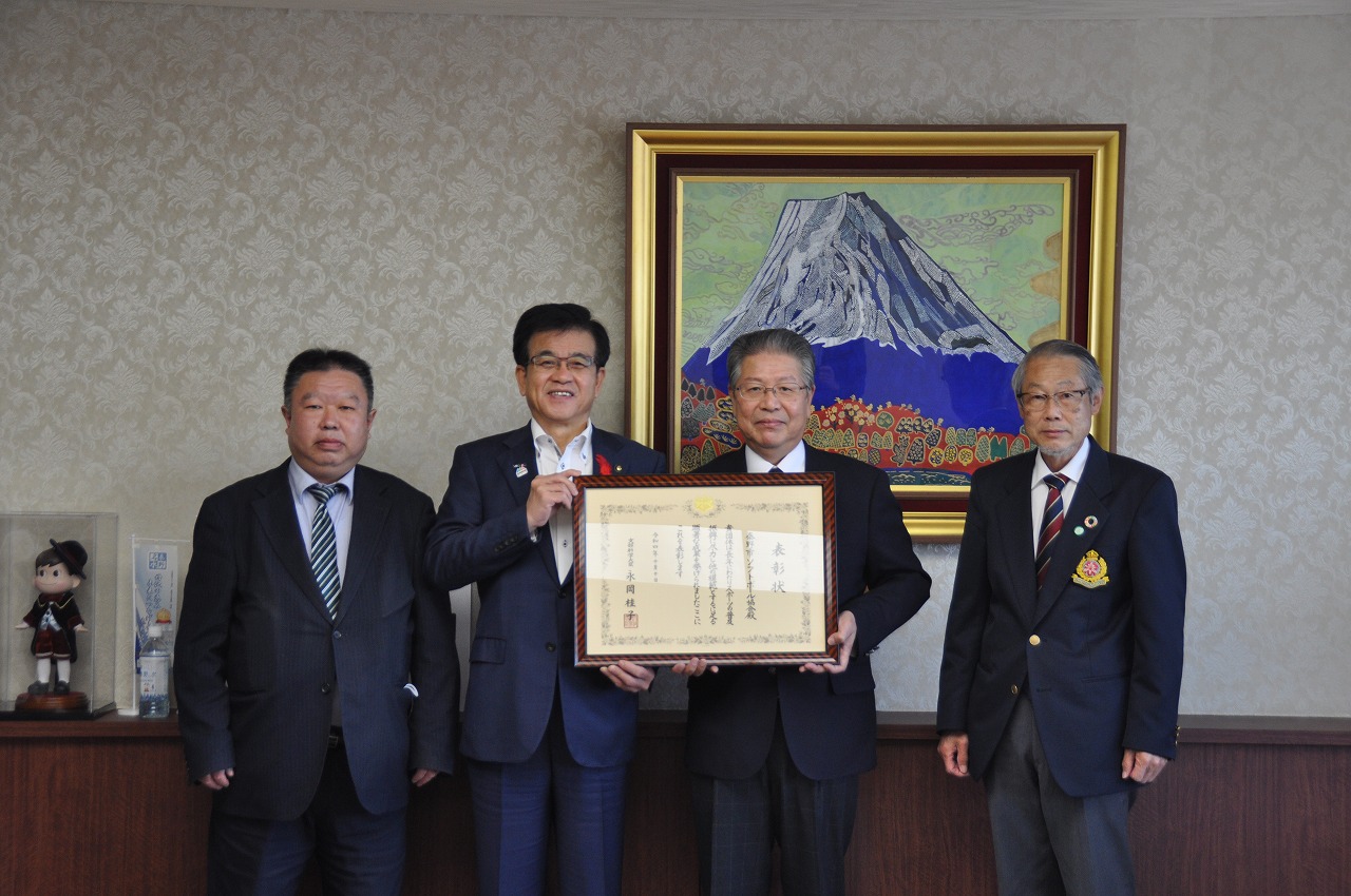スーツ姿の男性4人が横一列に並び、額に入った表彰状を中央で持ち、背景には富士山を描いた額縁の絵が飾られている記念撮影の写真