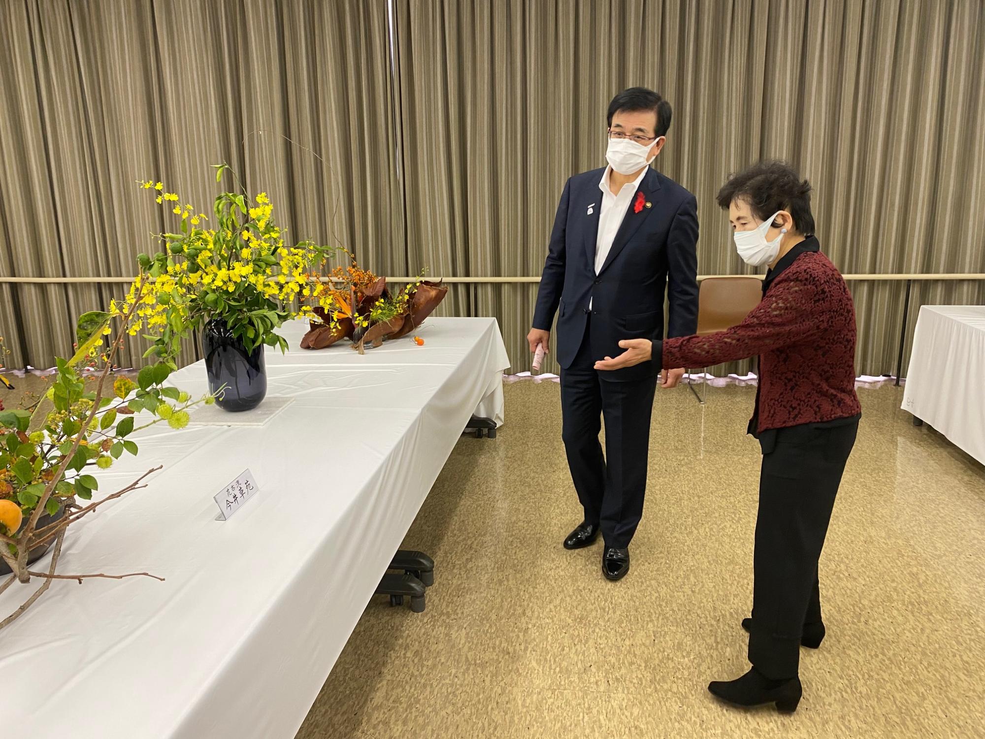 白いテーブルの上に黄色い花を生けた黒い花瓶が置かれ、スーツを着た男性に赤い上着を着た女性がその花を見ながら説明をしている様子の写真