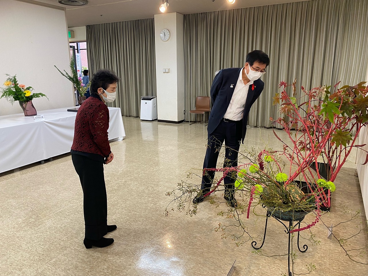 室内で、赤い枝と緑の花を使った生け花が飾られており、スーツ姿の男性が前かがみになって花を見つめ、女性と一緒に生け花作品を鑑賞している写真