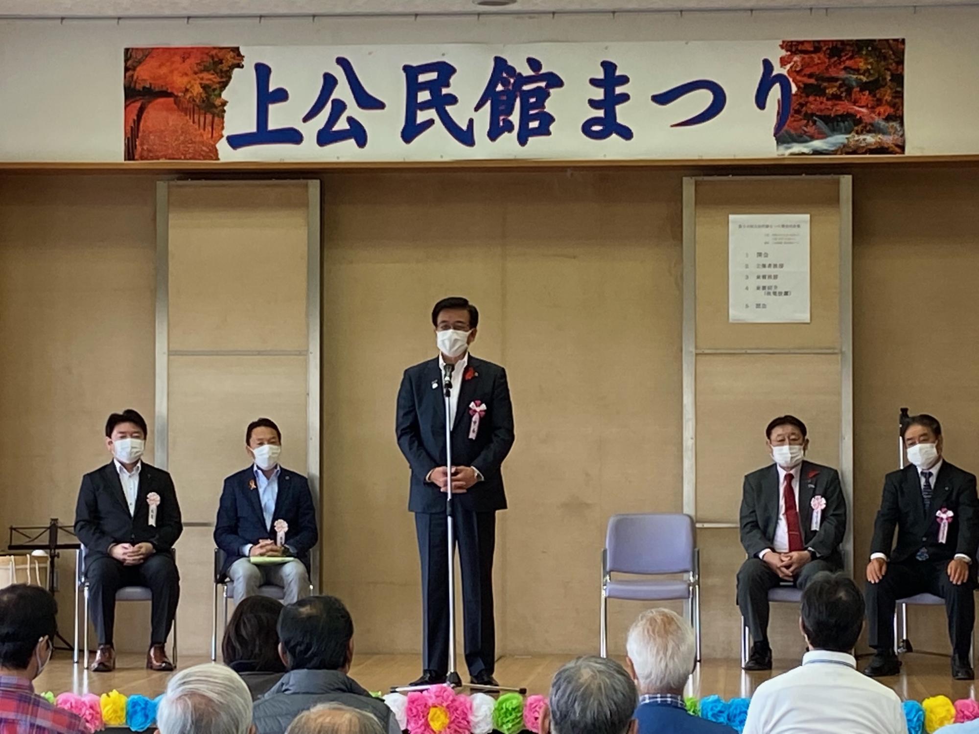「上公民館まつり」と書かれた横断幕の下で、スーツを着た男性がマイクの前に立ち、後ろに数人の来賓が座っており、観客が話を聞いている写真