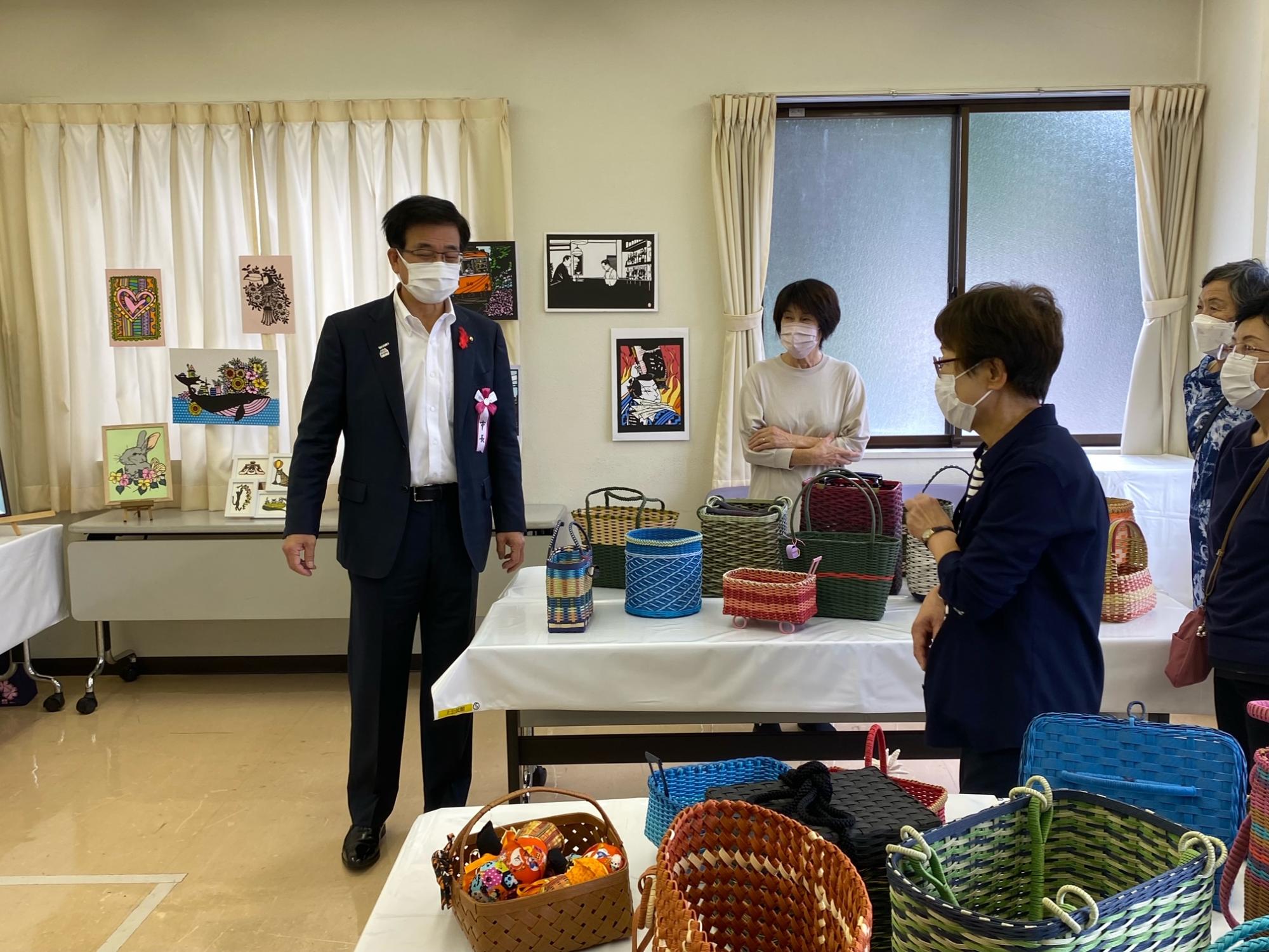 部屋の中に展示されたカラフルな手作りのカゴやバッグを、マスクをつけたスーツ姿の男性が鑑賞している写真