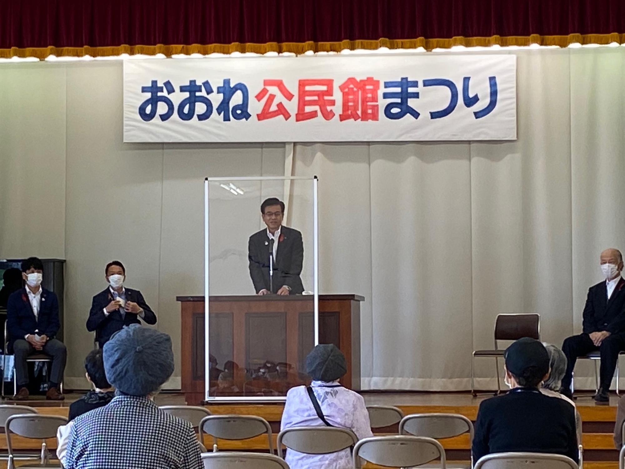 「おおね公民館まつり」と書かれた横断幕の下で、演台の後ろに立ったスーツ姿の男性が透明なアクリル板越しに話をしており、観客が椅子に座って聞いている写真