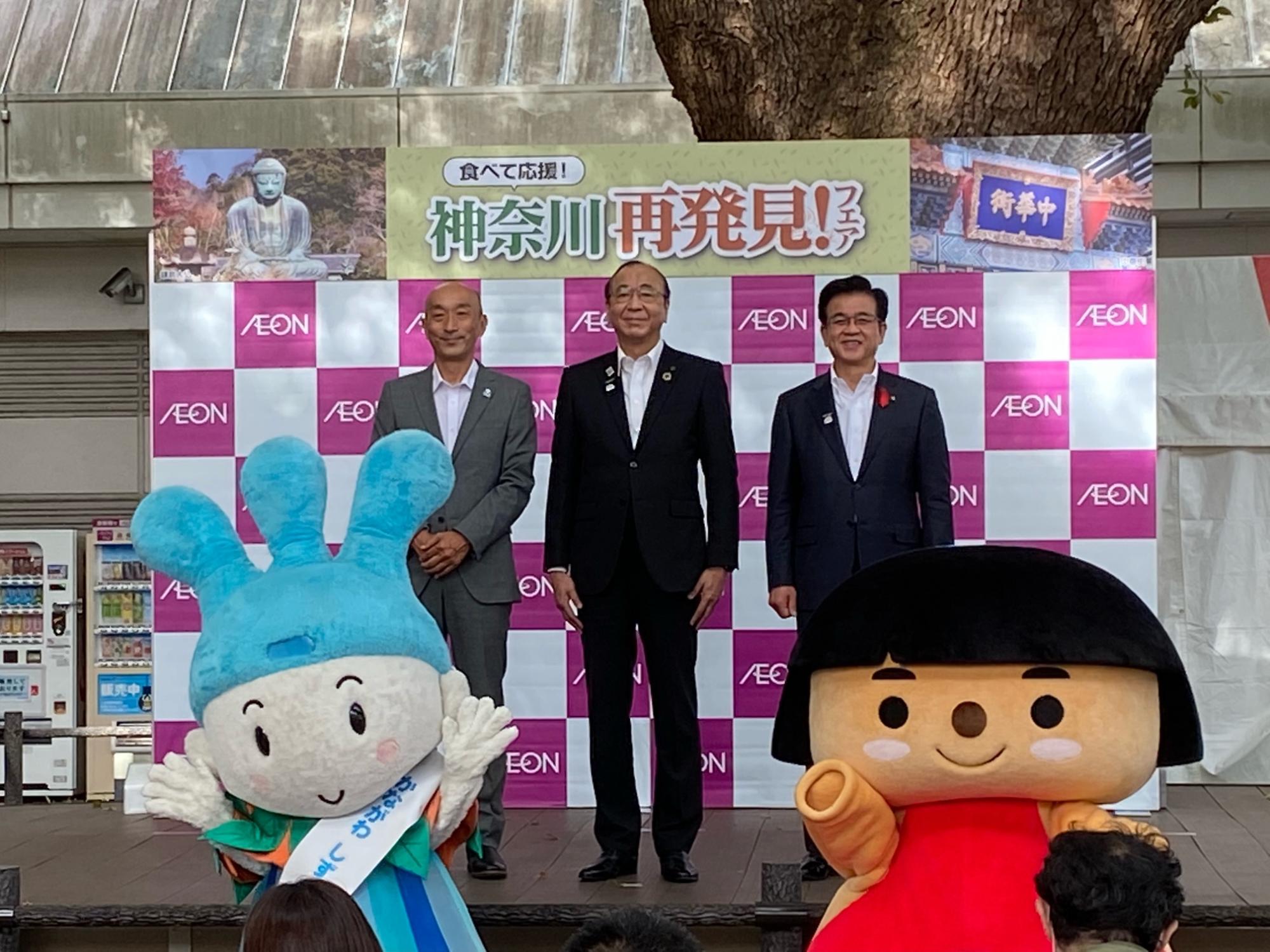 神奈川再発見の横断幕を背景に、スーツ姿の3人の男性が2体のゆるキャラと一緒に笑顔で並び、記念撮影をしている写真