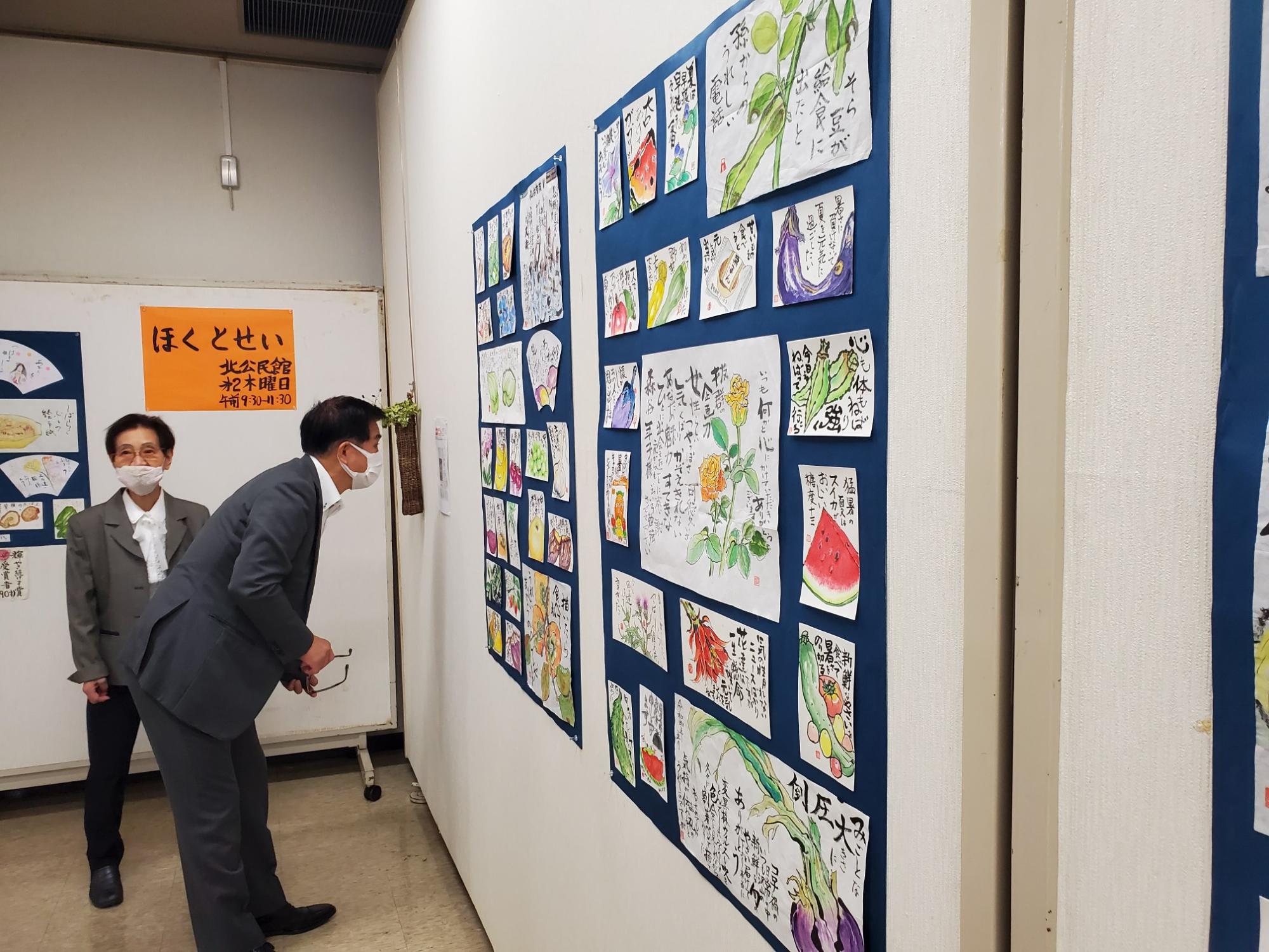 室内で展示されている野菜や花の絵手紙作品を、マスクをつけたスーツ姿の男性が身をかがめて熱心に見ている写真