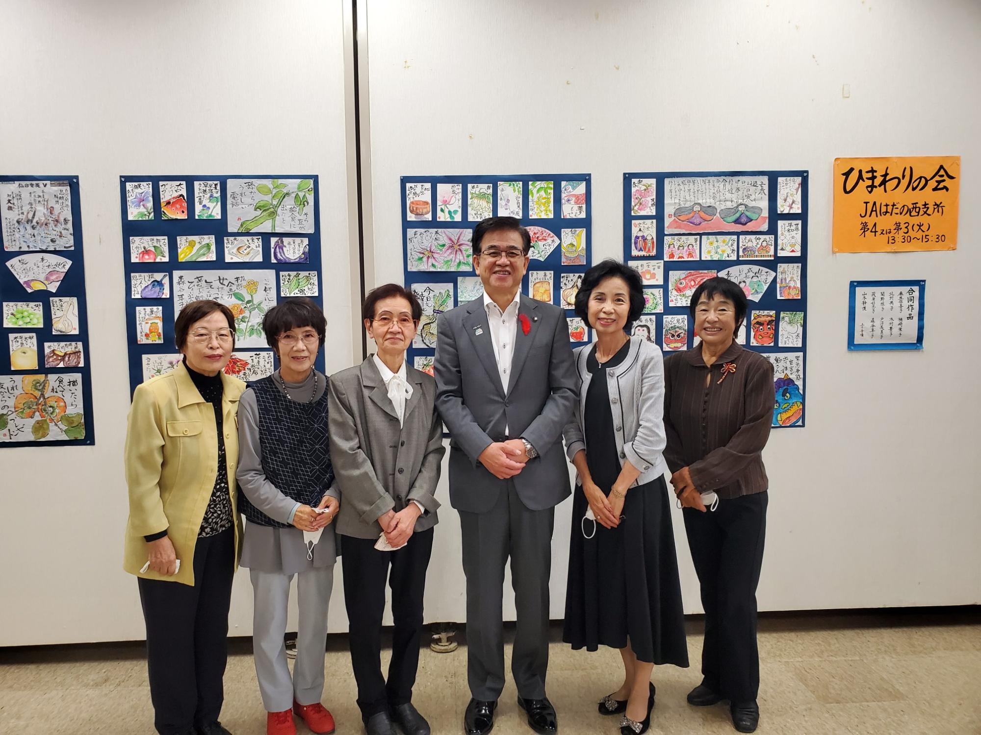 和風の絵手紙作品が壁に展示された部屋で、スーツ姿の男性が5人の女性たちと並んで笑顔で記念撮影をしている写真