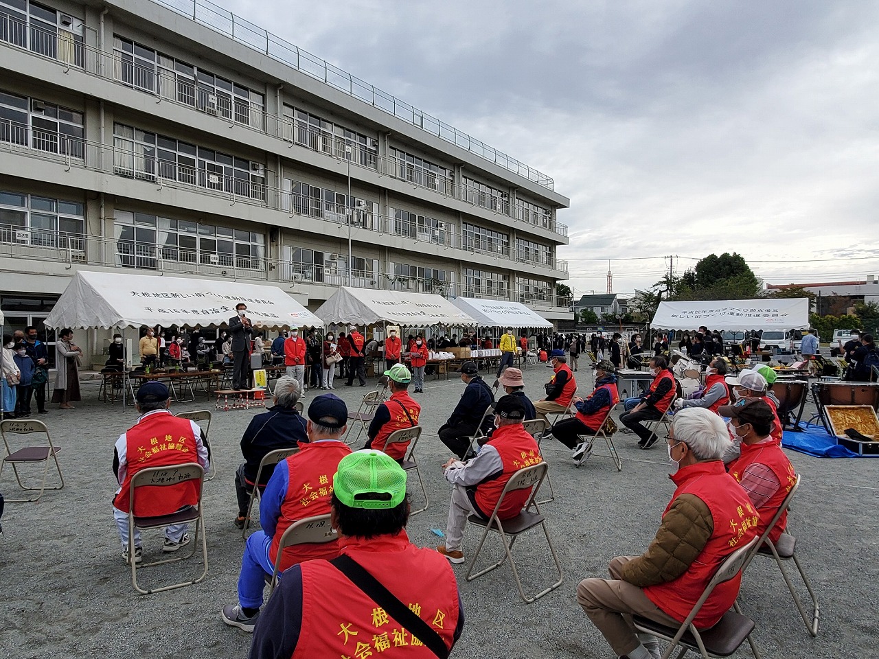 校庭に設営された複数のテントの前で多くの人が集まり、中央でスーツ姿の男性が演台に立ってマイクを持って話し、赤いベストを着た方が椅子に座って話を聞いている写真