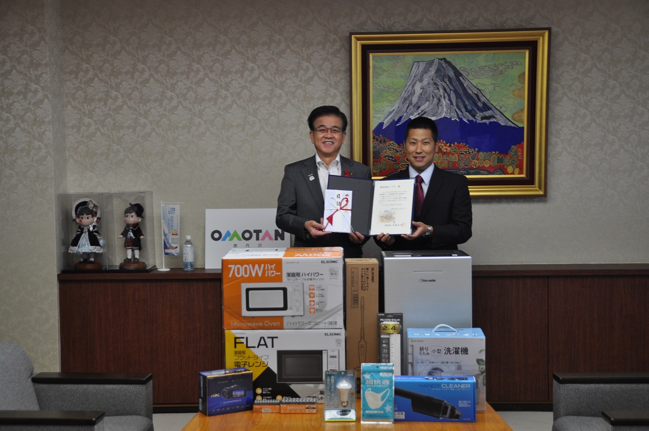 感謝状と目録を抱えた二人のスーツ姿の男性が、寄贈された家電製品や雑貨に囲まれて笑顔で立っている記念写真