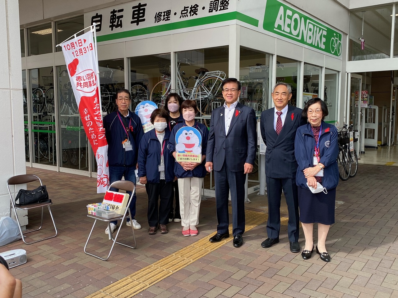 自転車売場の店頭で募金活動をしているグループと市長を含む7人が、赤い羽根共同募金の旗と鼻の赤い青いキャラクターの募金パネルの前で並んで記念撮影をしている写真