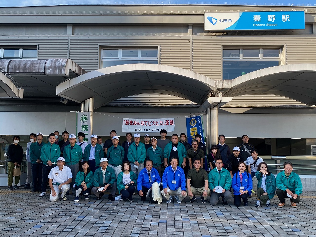 秦野駅前で「駅をみんなでピカピカ運動」の横断幕を掲げた大人数のグループが、緑や青のジャンパー姿で整列し記念撮影をしている駅前清掃活動の集合写真