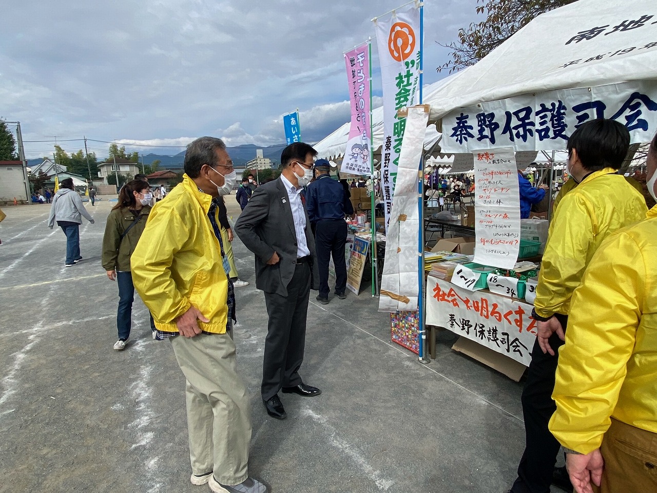 屋外のイベント会場で「秦野保護司会」の垂れ幕が掲げられた白いテントに、スーツを着た男性が黄色いジャンパーを着た数人のスタッフと話している写真