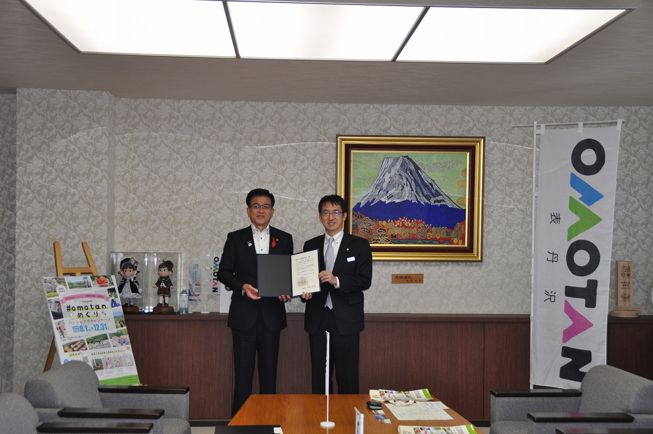 室内でスーツ姿の2人の男性が賞状を持って並んでおり、背後の壁には富士山のカラフルな絵画と「OMOTAN 表丹沢」と書かれたのぼりが飾られている受領式の記念撮影の写真