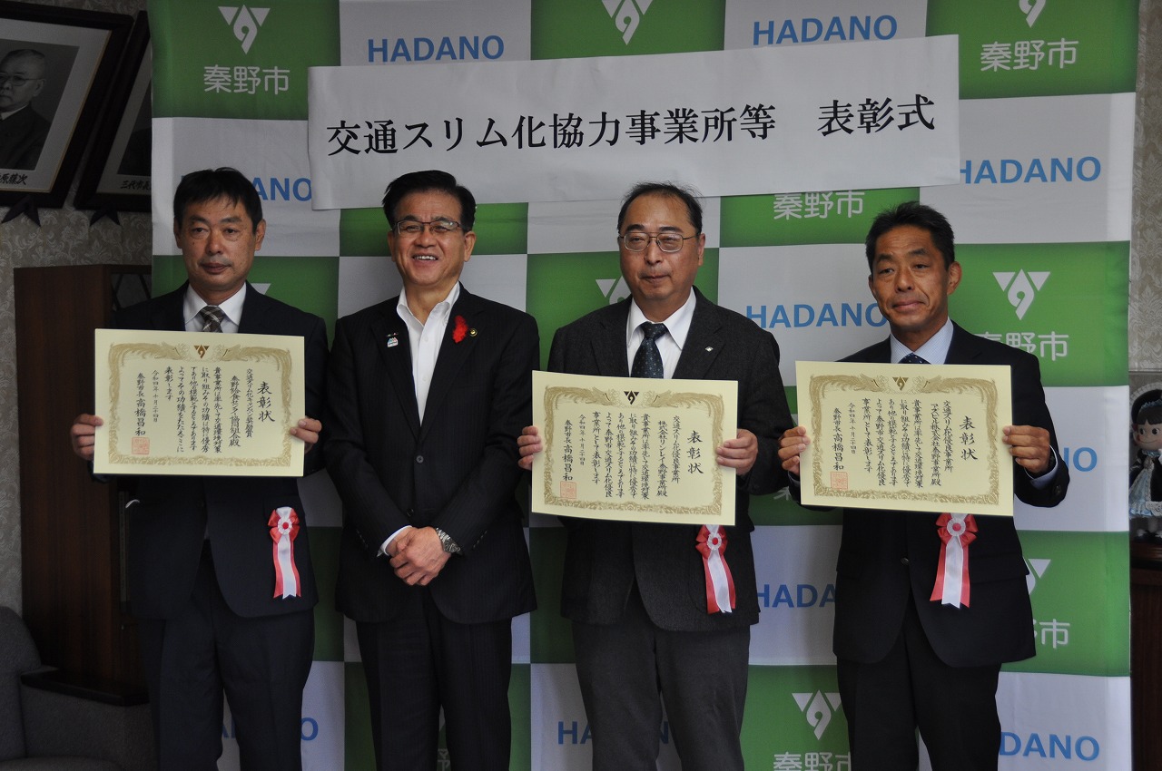 「交通スリム化協力事業所等 表彰式」と書かれている横断幕の前でスーツ姿の男性4人が並び、3人が表彰状を持っていている表彰式の記念撮影の写真