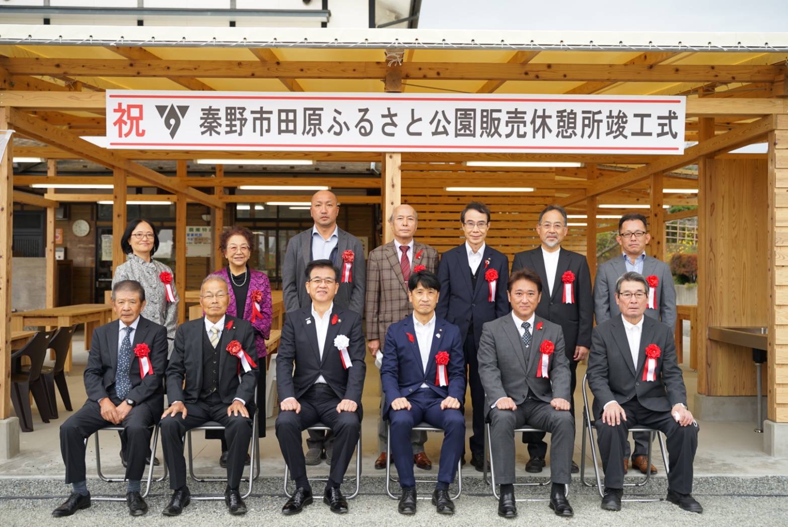「秦野市田原ふるさと公園販売休憩所竣工式」と書かれた横断幕の前に大勢の男女がスーツ姿で集まり記念撮影をしている写真