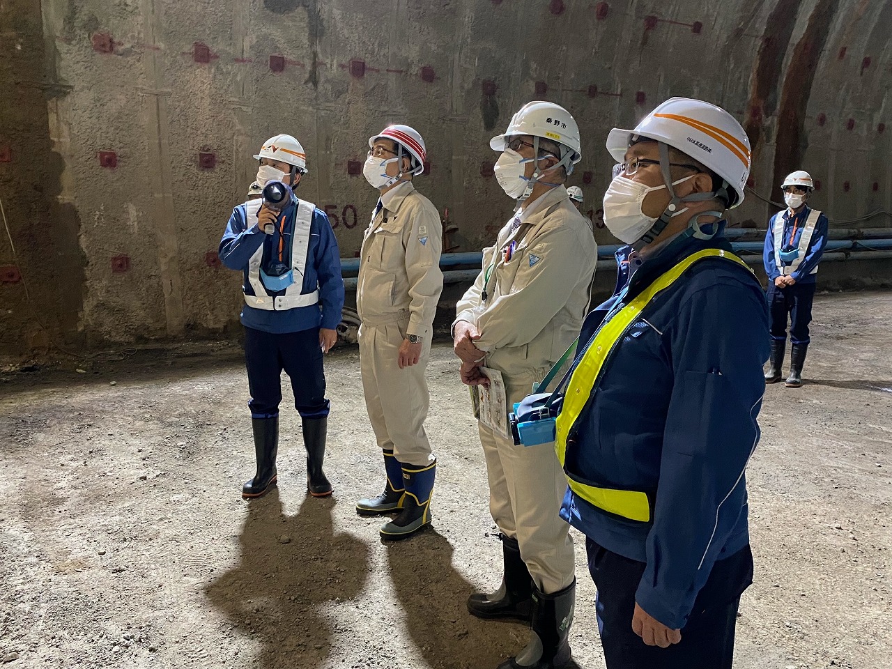 工事現場で、作業服にヘルメットとマスクを着用した市長と男性が並び、そのうちの一人が小型の拡声器を持ち話をしている写真