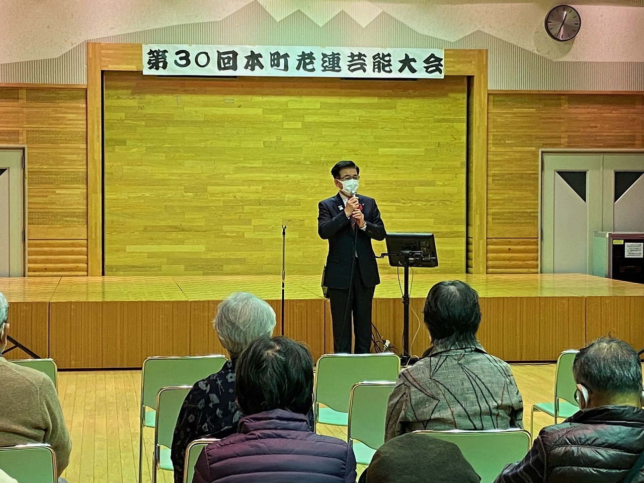 「第30回本町老連芸能大会」と書かれた横断幕の前でマスクを着用したスーツ姿の市長が、椅子に座った聴衆を前に話をしている写真