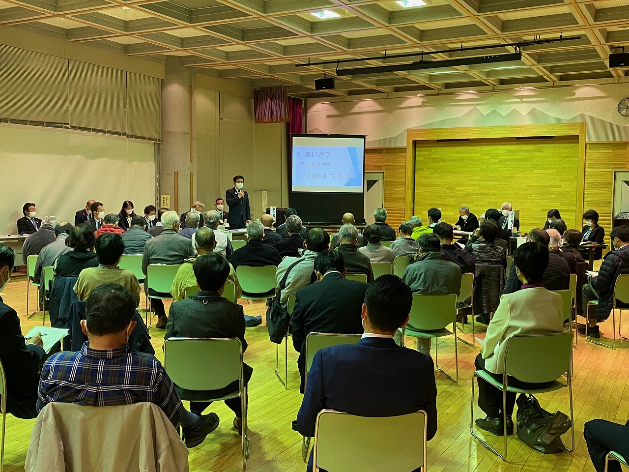 プロジェクターのスクリーンが設置された横に立ちマスクをつけたスーツ姿の市長が、多数の参加者に向かってマイクで話している様子の本町地区の市政懇談会の写真