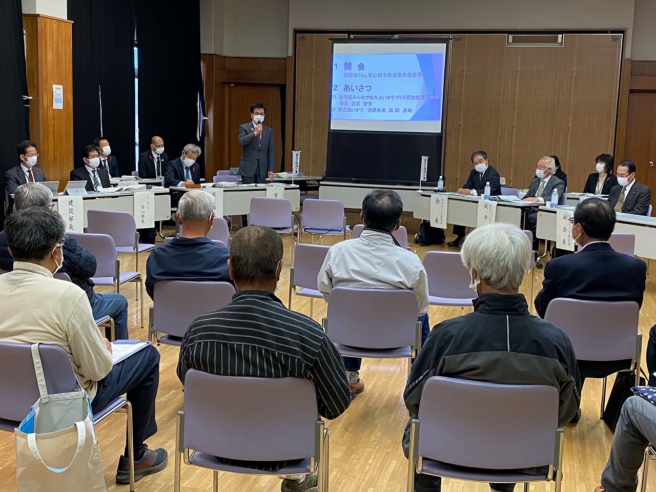椅子に座った多くの参加者と向かい合う形で並んだ会議室で、スーツ姿の市長がマイクを持って話している様子の上地区の市政懇談会の写真
