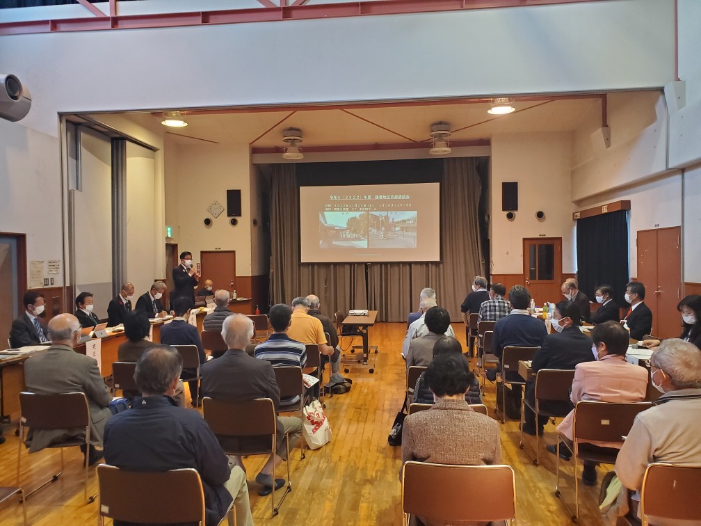 市長が、椅子に座った大勢の聴衆を前に、プロジェクターに映し出されたスライドを使って話をしている様子の鶴巻地区の市政懇談会の写真