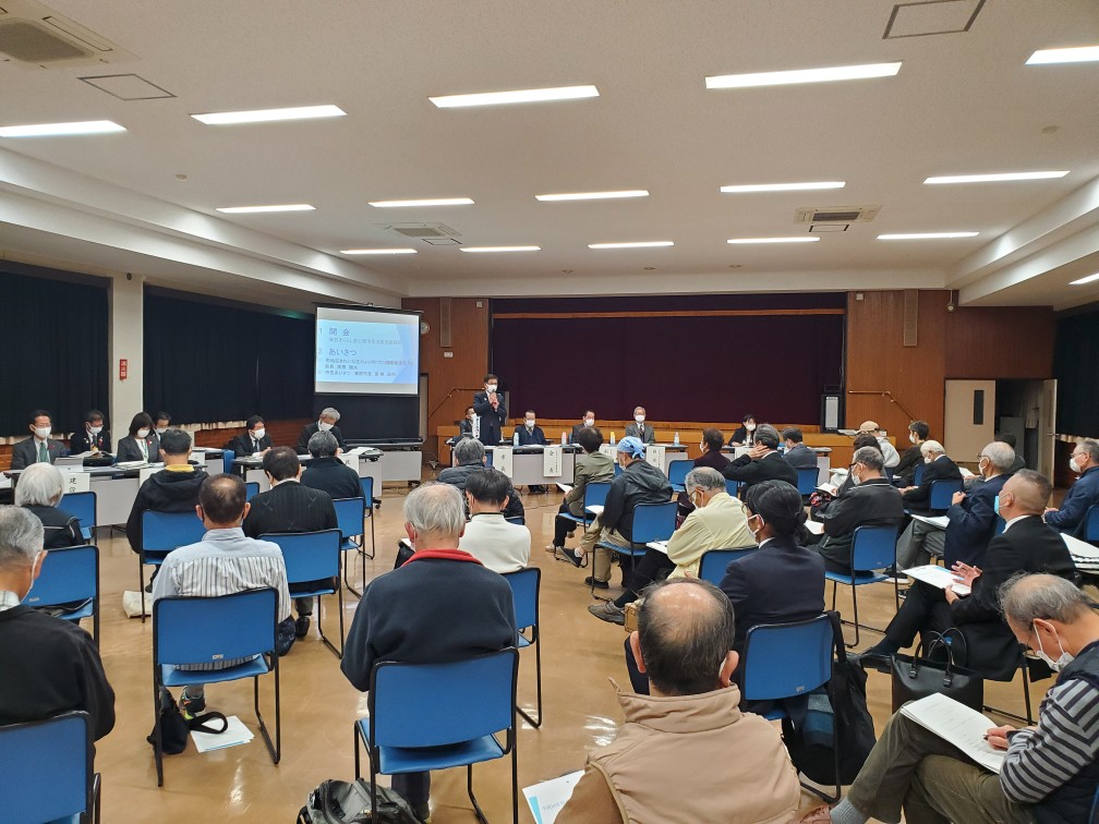 椅子に座った多くの参加者が集まる会議室で、プロジェクターの前に立つスーツ姿の市長がマイクで話をしている南地区の市政懇談会の写真