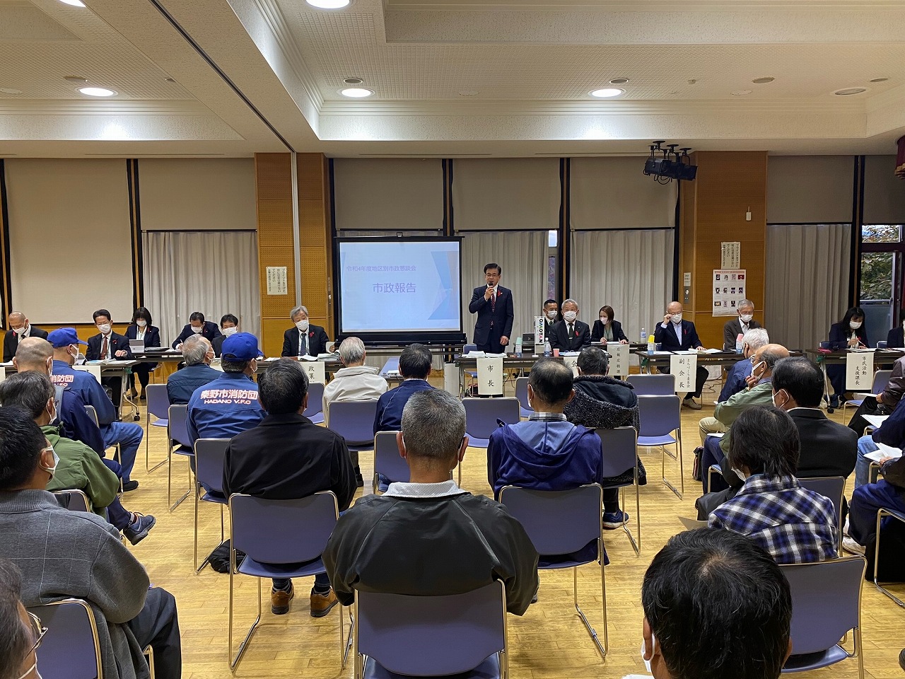「市政報告」と表示されたプロジェクターの前で、マスクをつけたスーツ姿の市長が、椅子に座った多くの人々を前に話をしている北地区の市政懇談会の写真