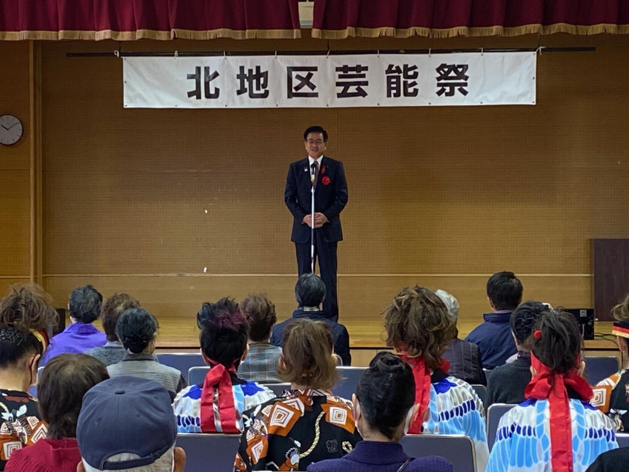「北地区芸能祭」と書かれた横断幕の舞台の中央でマスクを着用したスーツ姿の市長が、椅子に座った聴衆を前に話をしている写真