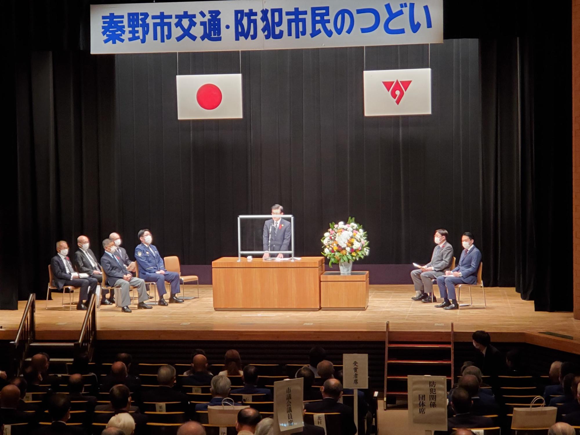 「秦野市交通・防犯市民のつどい」と書かれた横断幕が吊るされた体育館の壇上で、スーツ姿の市長がマイクを使って話をしており、周囲には警察官や関係者が並んで座っている写真